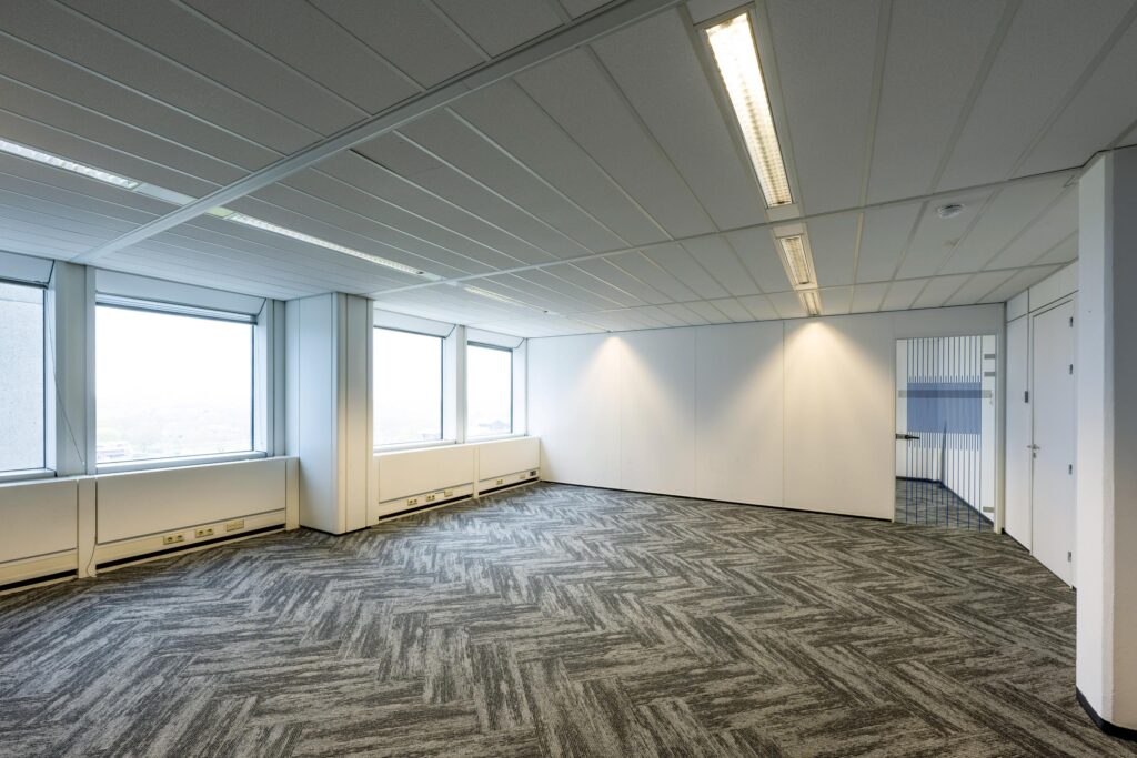 Empty office space at Laan van Kronenburg with large windows, patterned carpet, and white walls.
